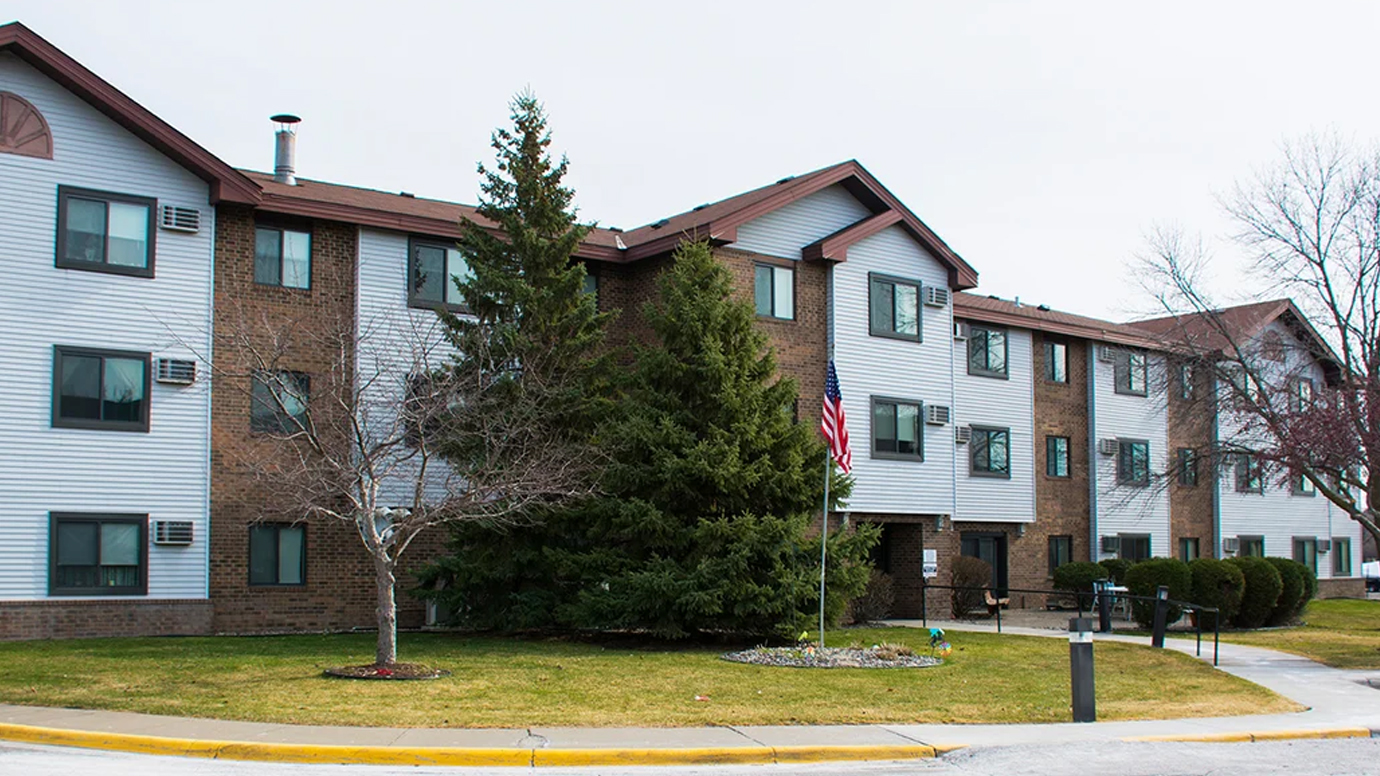 A three-story apartment building with landscaping and an American flag near the front door.